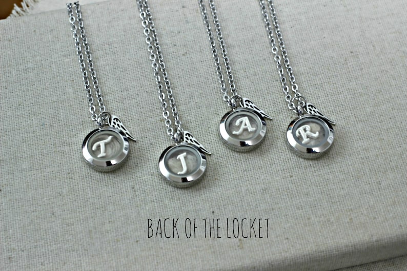 May include: Four silver locket necklaces with angel wings charms. Each locket has a different initial inside: T, J, A, and R. The back of the locket is visible in the image.
