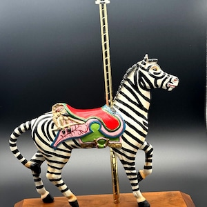 May include: A carousel zebra figurine with black and white stripes, a red, green, and pink saddle, and a gold-colored pole. It stands on a wooden base with a brass plaque reading PTC #6 CAROUSEL COLLECTION This Original ZEBRA is on the KIT CARSON COUNTY CAROUSEL - BURLINGTON, COLORADO #24 OF 250.