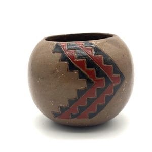 May include: A round, tan-colored ceramic pot with a wide, open top. The exterior surface features a prominent, hand-painted geometric design consisting of nested, stepped chevron patterns in alternating bands of deep black and dark red paint.