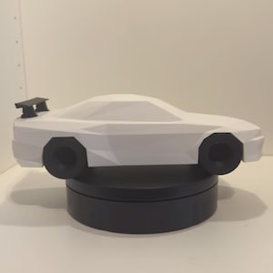 Könnte beinhalten: Ein weißes Low-Poly-Modell eines Sportwagens mit schwarzem Heckspoiler und schwarzen Rädern. Das Auto steht auf einem runden, schwarzen Präsentationsständer. Das Design besteht aus scharfen, geometrischen Facetten, die die Karosserieform definieren.