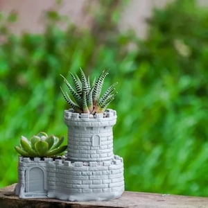 May include: A light gray, castle-shaped planter with a brick texture, featuring a tall central tower and a lower side section. Two green succulents are planted inside the structure, one in the tower and one in the lower section.
