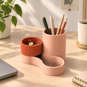 Op de afbeelding: Een bureau-organizer met meerdere vakken, bestaande uit een hoge, gladde, lichtroze cilindrische pennenhouder, een kortere, geribbelde, terracotta-kleurige ronde houder en een lage, gebogen, lichtroze schaal met zilveren paperclips.