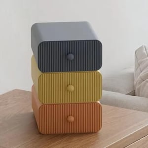 May include: A stack of three rectangular storage drawers with a vertical ribbed texture on the front panels. The top drawer is dark grey, the middle drawer is mustard yellow, and the bottom drawer is burnt orange, each with a small round knob.