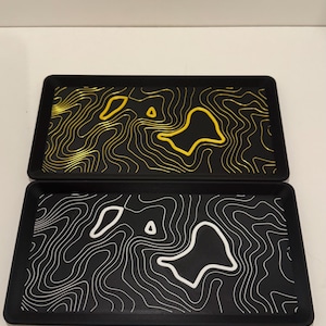 Könnte beinhalten: Zwei rechteckige schwarze Tabletts mit topographischem Kartenmuster. Das obere Tablett zeigt goldene Höhenlinien, während das untere Tablett weiße Höhenlinien aufweist. Beide Tabletts haben erhöhte Ränder und eine flache Basis mit matter Oberfläche.