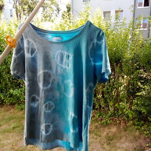 Puede incluir: Una camiseta de manga corta con un patrón tie-dye en tonos azules y grises. La prenda tiene un cuello redondo y muestra una serie de motivos circulares con bordes blancos dispersos por la tela, que pasan de un gris azulado oscuro a la izquierda a un azul turquesa brillante y sólido en el lado derecho.