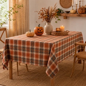 May include: A rectangular tablecloth featuring a plaid pattern in shades of orange, brown, and cream. The fabric drapes over a table, which is decorated with a carved jack-o'-lantern, a small pumpkin, a vase of dried flowers, and a candle.