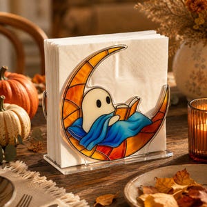 Peut inclure: Un porte-serviettes en acrylique transparent avec un insert décoratif style vitrail représentant un fantôme blanc lisant un livre sur un croissant de lune. La lune est orange et jaune, et le fantôme est enveloppé dans une couverture bleue.