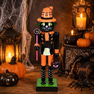 May include: A wooden nutcracker shaped like a black cat wearing an orange witch hat. The figure features green eyes, a pink lollipop in one hand, and a small black pumpkin bucket labeled BOO in the other. It stands on a green rectangular base.