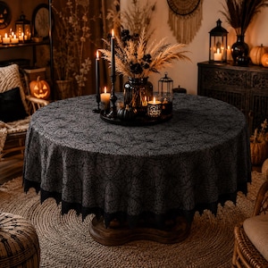 May include: A round black tablecloth featuring a repeating spiderweb pattern across the fabric, finished with a scalloped edge. The tablecloth is draped over a circular table, serving as a base for a centrepiece of candles and dried grasses.
