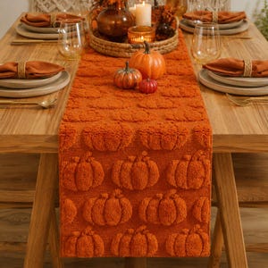 May include: A burnt orange table runner featuring a repeating tufted pumpkin pattern, displayed on a wooden table with place settings including orange napkins, clear glasses, and a centerpiece of three pumpkins in a woven tray.