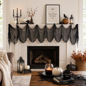May include: A black cheesecloth fabric draped across a fireplace mantel, decorated with white and orange pumpkins, a black candelabra, a framed sign reading A HAUNTING WE WILL GO, a black lantern, and a crow figurine, with books and candles below.