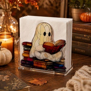Peut inclure: Un porte-serviettes en acrylique transparent orné d'une illustration imprimée d'un fantôme crème aux yeux noirs assis sur une pile de livres colorés, lisant un livre rouge ouvert. Le support contient une pile de serviettes en papier blanc.