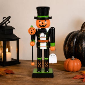Könnte beinhalten: Eine dekorative Nussknacker-Figur mit einem geschnitzten, orangefarbenen Kürbiskopf als Jack-o'-Lantern, die einen schwarzen Zylinder und eine schwarze Fliege trägt. Die Figur hält einen Stab mit Kürbisaufsatz und einen weißen, geisterförmigen Eimer.