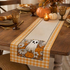 Op de afbeelding: Een rechthoekige tafelloper met een crèmekleurig middenpaneel voorzien van een print van een witte geest, oranje pompoenen en witte madeliefjes. De loper heeft een geel-witte gingham-rand langs de lange zijden.