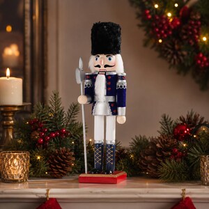 May include: A wooden nutcracker figurine wearing a tall black fuzzy hat, a blue jacket with silver accents, and white trousers. The figure has a white beard, a mustache, and holds a silver-colored staff while standing on a red rectangular base.