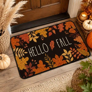 May include: A rectangular black doormat featuring the white text HELLO FALL in the center, surrounded by a border of printed autumn leaves in shades of orange, yellow, and brown. The mat has rounded corners and a black trim along the edges.