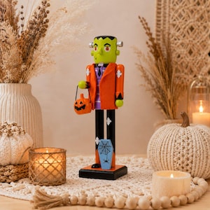 May include: A Frankenstein-themed nutcracker figure with green skin, a bright orange jacket, and black trousers. The figure holds a small orange jack-o'-lantern bucket and features a light blue coffin-shaped accent with a black spiderweb design.