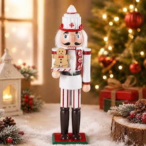 Op de afbeelding: Een decoratieve notenkrakerfiguur in een wit en rood uniform met pepermuntmotief en bijpassende hoed. Het figuur houdt een klein peperkoekmannetje op een dienblad vast en heeft wit haar, een zwarte snor en donkerbruine laarzen.