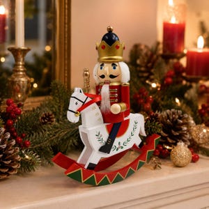 Könnte beinhalten: Eine hölzerne Nussknacker-Figur mit goldener Krone mit roten Akzenten und roter Uniform, montiert auf einem weißen Schaukelpferd. Das Pferd zeigt grün-rote geometrische Muster an den Kufen und zarte grüne Blattmotive an den Seiten.