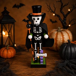 May include: A skeleton nutcracker wearing a black top hat with an orange band and a black cape. The figure holds a staff topped with a bat and stands on a green base with a sign that reads Ghost Town. The skeleton features white bone details.