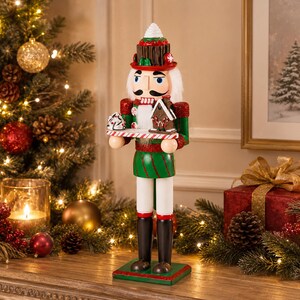 Peut inclure: Une figurine de casse-noisette portant un uniforme vert et rouge avec un chapeau en forme de gâteau au chocolat. La figurine tient un plateau avec une petite maison en pain d'épice et un cupcake givré blanc. Cheveux blancs et bottes.