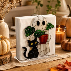 May include: A clear acrylic napkin holder featuring a decorative front panel with a stained glass style illustration of a white ghost holding a potted monstera plant, accompanied by a black cat with yellow eyes sitting at the base of the ghost.