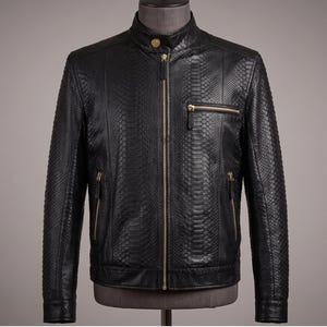 May include: A black jacket with a textured snakeskin pattern, featuring a stand-up collar with a gold button, a central gold zip, and three gold zipped pockets on the front. The garment has long sleeves and a straight hemline design.