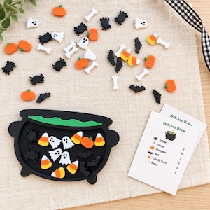 Peut inclure: Un plateau noir en forme de chaudron contenant de petites pièces en résine sur le thème d'Halloween, dont des fantômes blancs, des citrouilles orange, des araignées noires, des chauves-souris, des os blancs et des bonbons maïs.