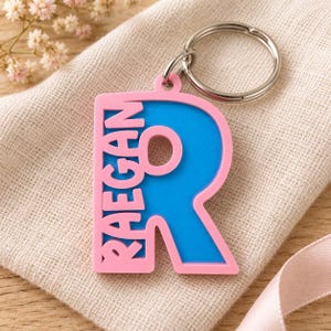 May include: A custom keychain featuring a large, bright blue letter R with a pink border. The name RAEGAN is vertically embossed in pink letters along the left side of the letter R. A silver metal split ring is attached to the top of the keychain.