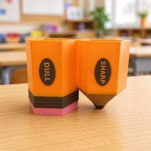 Op de afbeelding: Een oranje pennenhouder met twee vakken in de vorm van een potlood. De ene kant heeft een bruine basis met een roze gum en een ovaal label met de tekst DULL. De andere kant heeft een bruine punt en een ovaal label met de tekst SHARP.