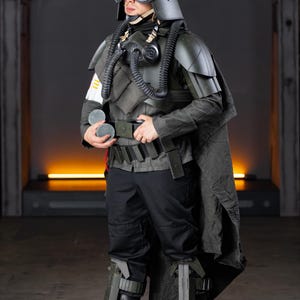 May include: A dark olive green and black tactical costume featuring a helmet with goggles, a chest piece with dual breathing tubes, shoulder armor, a long cape, a utility belt, and leg guards. The outfit includes black trousers and boots.