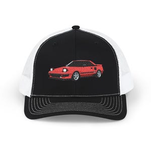 May include: A black and white trucker cap featuring an embroidered graphic of a red sports car on the front panel. The cap has a solid black front, a white mesh back, and a black brim with detailed stitching across the top of the visor.