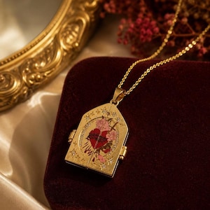 May include: A gold-toned locket necklace featuring a pointed top and a hinged design. The front displays a central illustration of a red heart surrounded by pink flowers, accented with small clear stones and star-shaped engravings on the metal surface.