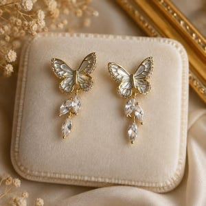 May include: A pair of gold-tone butterfly earrings featuring white enamel wings with clear crystal accents along the edges. Each earring includes a dangling cluster of three marquise-cut clear crystals suspended from the base of the butterfly.