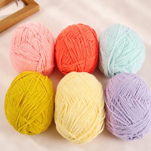 May include: Six round balls of soft, thick yarn arranged in two rows. The colours include light pink, bright orange, pale mint green, mustard yellow, cream, and light purple. Each ball features a textured, cord-like appearance with a consistent twist.