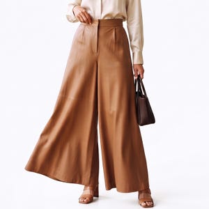 May include: High-waisted wide-leg trousers in a solid tan color, featuring a smooth finish, a structured waistband with a front zipper closure, and a clean, flowing silhouette that extends to the floor, paired with a light beige long-sleeve shirt.