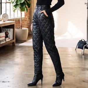 May include: High-waisted black trousers featuring a quilted diamond pattern throughout the fabric. The pants have a structured waistband with a single button closure, side pockets, and a tapered leg silhouette, paired with black pointed-toe boots.