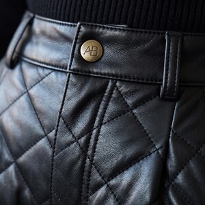 May include: A black quilted leather skirt featuring a high waistband with belt loops and a central brass-toned button embossed with the letters AB. The garment displays a diamond-shaped stitched pattern across the surface of the leather material.