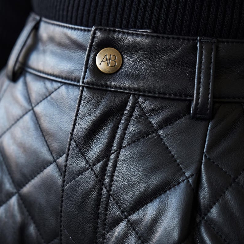 May include: A black quilted leather skirt featuring a high waistband with belt loops and a central brass-toned button embossed with the letters AB. The garment displays a diamond-shaped stitched pattern across the surface of the leather material.