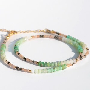 May include: A delicate beaded bracelet featuring small, faceted mint green, pale peach, and dark metallic gray beads arranged in a repeating pattern. The strand is accented with tiny gold-toned metal beads and includes a gold-toned chain extender.