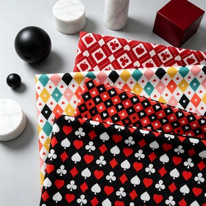 Op de afbeelding: Vier gevouwen stukken stof met patronen van speelkaarten. De bovenste laag is rood en wit met schoppen en klaveren. De tweede heeft een veelkleurig ruitpatroon. De derde is rood en zwart met kleine symbolen. De onderste laag is zwart met rode harten en witte schoppen en klaveren.