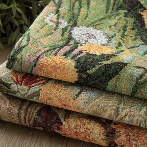 Op de afbeelding: Drie gevouwen lagen geweven stof met een dicht botanisch patroon. Het textiel vertoont een levendige mix van groene, gele, oranje en witte draden, wat een gedetailleerd tafereel van diverse bloemen en bladeren met een textuur creëert.