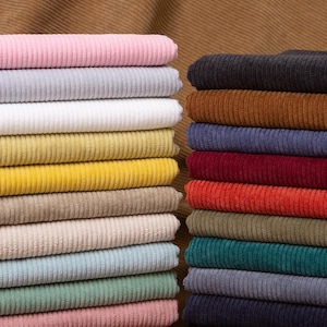 May include: Two stacks of folded corduroy fabric in a wide variety of colors including pink, light gray, white, yellow, beige, light blue, sage green, dark brown, burgundy, red, olive green, teal, navy blue, and black, all featuring a ribbed texture.