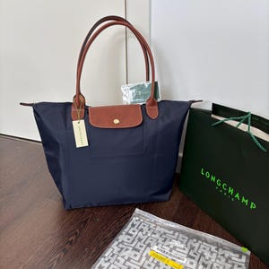 May include: A navy blue nylon tote bag with brown leather handles and a matching brown leather flap featuring a gold-tone snap button. A beige Longchamp brand tag is attached to the handle, and a dark green shopping bag with the text LONGCHAMP PARIS is nearby.