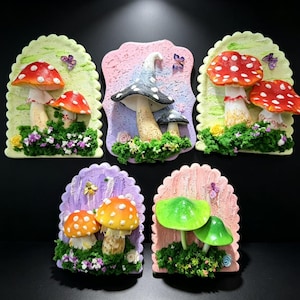 Puede incluir: Cinco placas decorativas de pared con setas esculpidas en varios colores, incluyendo rojo, azul, naranja y verde. Cada placa tiene un fondo texturizado, musgo artificial, flores pequeñas y detalles de insectos como mariposas y una abeja.