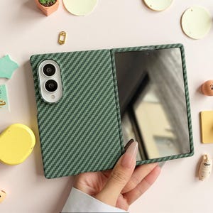 May include: A foldable phone case featuring a dark green carbon fiber pattern. The case has a rectangular camera cutout with a white border housing two circular lenses. The interior of the open case displays a reflective screen surface.