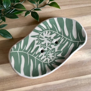 Puede incluir: Un jabonera de cerámica verde y blanca con un diseño de hojas. La jabonera tiene dos agujeros de drenaje.