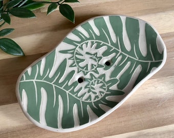 Könnte beinhalten: Eine grüne und weiße Keramik-Seifenschale mit einem Blattdesign. Die Schale hat zwei Abflusslöcher.