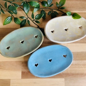 Puede incluir: Tres jaboneras de cerámica con recortes en forma de corazón. Las jaboneras son en tonos de azul, verde y crema.