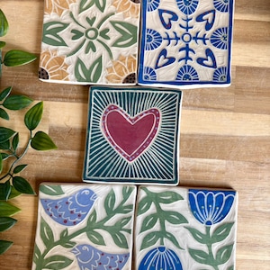 May include: Five square ceramic tiles featuring hand-painted designs. One tile has yellow and green floral patterns, another has blue and white heart motifs, one displays a central red heart, one shows two blue birds, and one features blue flowers.